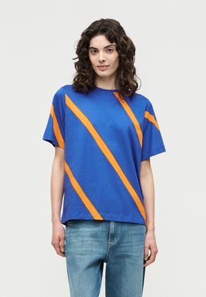 Mujer con cabello rizado oscuro que lleva una camiseta azul de manga corta con rayas diagonales naranjas y jeans azul claro, de pie contra un fondo liso.