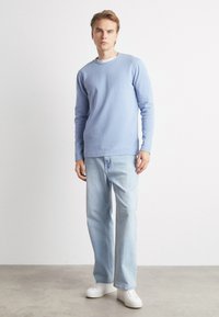 Camicia azzurra a maniche lunghe con scollatura rotonda, indossata sopra una maglietta bianca, abbinata a jeans azzurri e sneaker bianche.