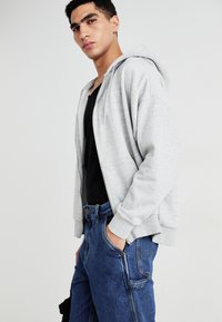 Light grey zip-up hoodie made of soft fabric, featuring a relaxed fit and side pockets, worn over a black shirt and blue jeans.