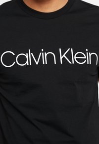 Černé bavlněné tričko s kulatým výstřihem, na kterém je na hrudi vytištěn bílý nápis "Calvin Klein" v moderním písmu bez serifa.