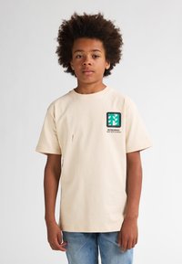 Beige katoenen t-shirt met korte mouwen, voorzien van een groen en zwart grafisch logo op de borst en een kleine rode vlek nabij de zoom.