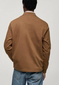 Hnědá mikina s jemnou texturou, disponující límečkem, dlouhými rukávy a volným střihem. Nositá s světlými modrými džíny.