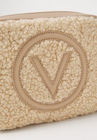 Μπεζ τσάντα με υφή, με μαλακή και αφράτη επιφάνεια, που διαθέτει ένα ξεχωριστό κυκλικό λογότυπο με "V" στο κέντρο, περιτριγυρισμένο από λείο δέρμα.