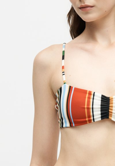 Gros plan d'une femme portant un haut de bikini rayé multicolore avec des bretelles fines sur un fond blanc uni.