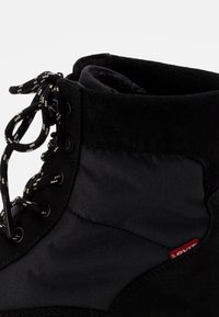 Botín negro de ante y tela, con cordones negros y una superficie texturizada. Incluye una pequeña etiqueta roja de Levi's en el lateral.