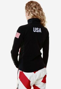 Giacca nera con zip che presenta "USA" in bianco sul retro e una toppa della bandiera americana sulla manica sinistra. Tessuto testurizzato con un design aderente.