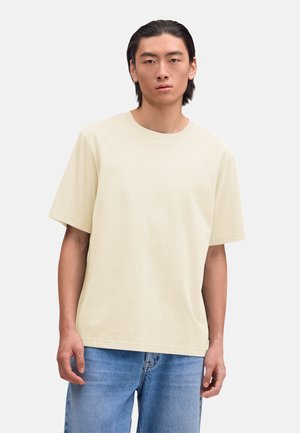 Young man with straight dark hair wearing a plain light beige short-sleeve T-shirt and blue denim jeans against a white background.