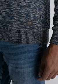 Gebreide marineblauwe en grijze trui met een textuurpatroon, geribbelde manchetten en een genaaid etiket aan de zoom. Gepaard met blauwe denim jeans.