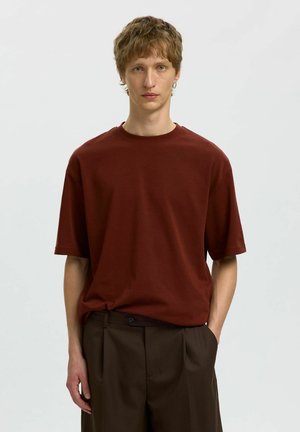Jeune homme aux cheveux courts châtain clair portant un t-shirt ample bordeaux et un pantalon marron foncé, debout avec une main dans la poche.