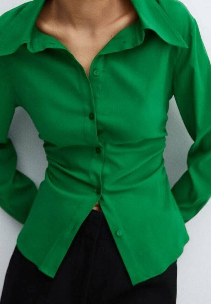 Femme portant une chemise ajustée verte vif à boutons avec un large col et un pantalon noir, les mains reposant derrière son dos.