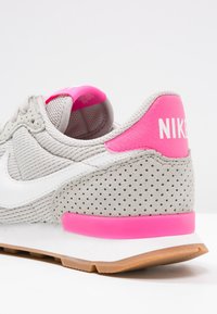 Harmaa Nike-tenniskenkä vaaleanpunaisilla yksityiskohdilla, valkoisella swooshilla ja reikäistetuilla nahkayksityiskohdilla valkoisella pinnalla.
