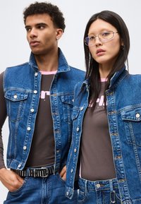Deux mannequins portent des vestes en jean bleu assorties sur des hauts gris avec des cols roses, associées à des jeans bleus, tournant légèrement le dos à la caméra.