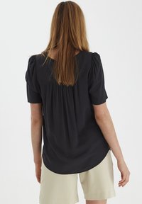 Blusa nera a maniche corte con pannello posteriore arricciato e orlo curvo, abbinata a pantaloncini beige chiaro. Tessuto morbido.