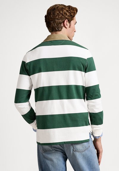 Vīrietis ar cirtainiem brūniem matiem, tērpies zaļi baltā svītrainā garroku regbija kreklā un gaiši zilās džinsās, skatās prom no kameras.