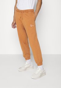 Orange sweatpants with an elastic waistband and cuffs, featuring a white embroidered logo on the left thigh. Worn with light-colored sneakers.