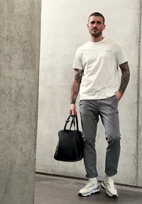 Homme aux bras tatoués portant un t-shirt blanc et un pantalon gris, tenant un sac en cuir noir, debout devant un mur en béton.