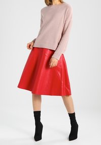 Kvinna i en ljusrosa tröja, röd A-linjeformad läderkjol och svarta klackade ankelskons, som står mot vit bakgrund.