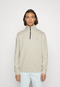 Felpa beige a maniche lunghe con colletto alto e zip a quarti. Presenta polsini e orlo a coste, con un logo ricamato sul petto. Texture liscia.