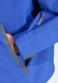 Modrá nepremokavá bunda s hladkým povrchom. Obsahuje reflexné detaily, nastaviteľné manžety a vrecko integrované do dizajnu.