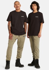 Svarte bomulls t-skjorter med Timberland-logo, kombinert med olivengrønne joggebukser med merkevare, og svarte snørestøvler med teksturerte såler.