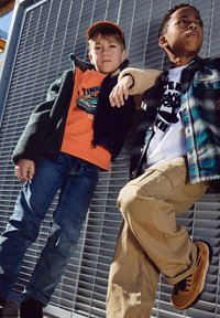 Due bambini posano contro una griglia metallica. Uno indossa un felpa arancione e una giacca in pile verde scuro, l'altro una camicia a quadri e pantaloni color kaki.
