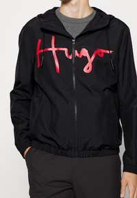 Black windbreaker jacket with a zip front, featuring a pink-to-red gradient "Hugo" logo on the chest. Includes a hood and elastic cuffs.