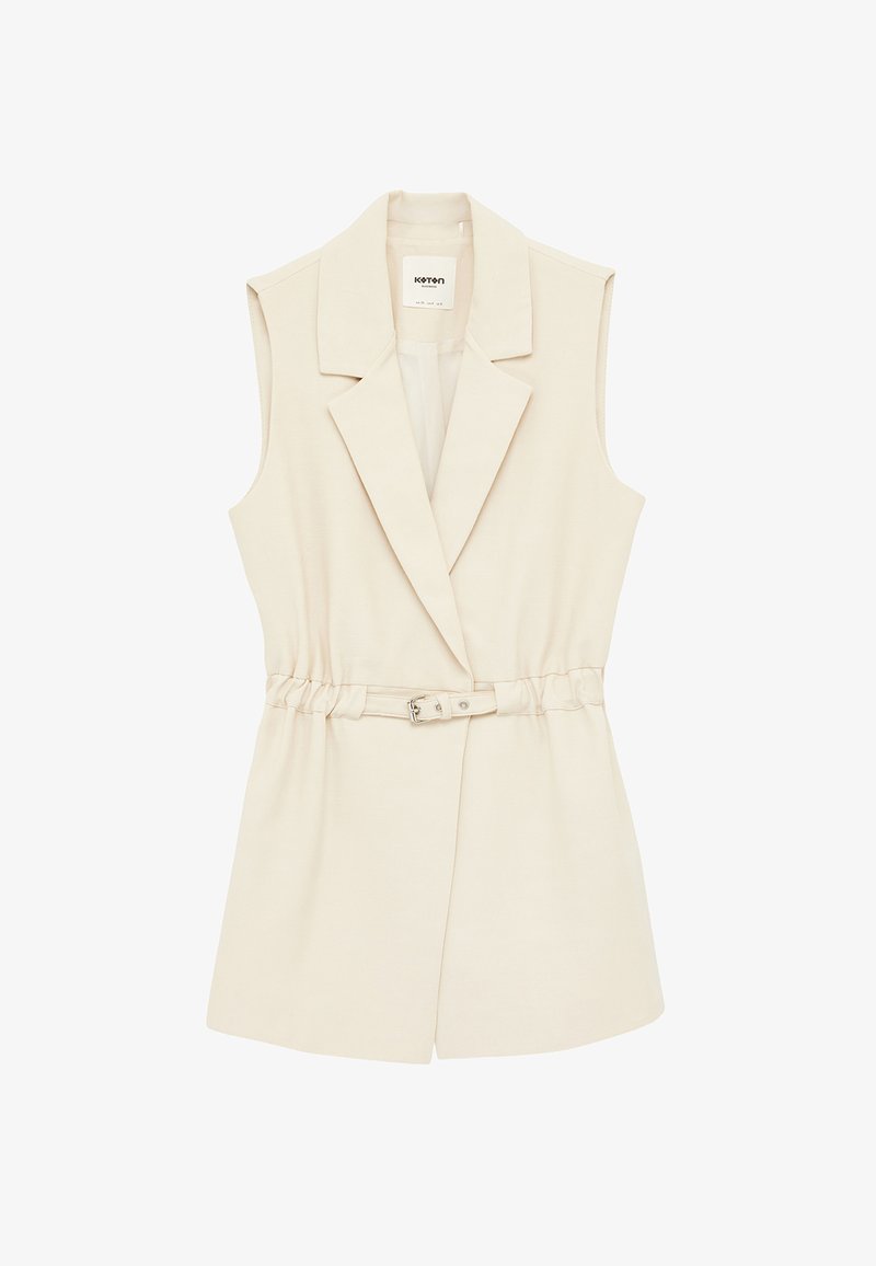 Veste sans manches beige à larges revers et une ceinture assortie avec une boucle métallique à la taille.