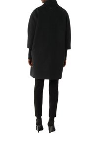 Cappotto di lana nero con un colletto alto e maniche ampie, caratterizzato da una texture liscia e un design minimalista, abbinato a pantaloni neri aderenti e tacchi.