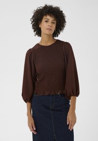 Haut marron texturé avec manches bouffantes, présentant un design froncé et un ourlet à volants. Associé à une jupe en denim foncé.
