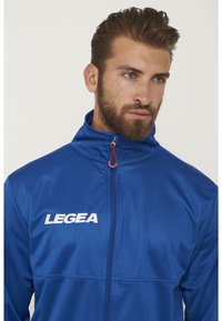 Giacca atletica blu con colletto alto, chiusura frontale a metà zip e logo "LEGEA" bianco. Tessuto sintetico liscio con una finitura leggermente lucida.