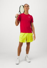 Rød idretts T-skjorte med logo, paret med lyse gule shorts med lommer. Personen holder en tennisracket.