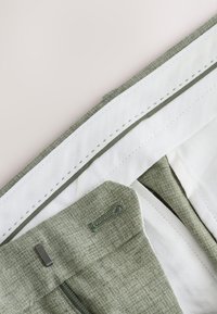 Pantalon en tissu vert clair avec une finition texturée, doublure blanche, surpiqûres vertes contrastées et un petit accent métallique à la taille.