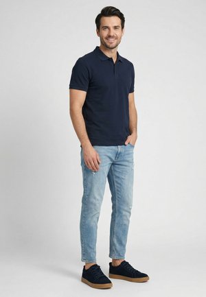Muž stojící, oblečený v tmavě modrém polo tričku, světle modrých opraných džínách a černých teniskách s gumovou podrážkou, usmívající se do kamery.