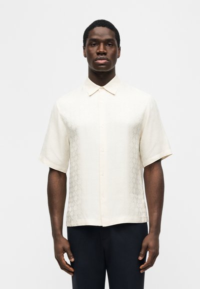 Άνδρας που φοράει πουκάμισο με κοντό μανίκι σε κρεμ χρώμα με διακριτικά σχέδια και μαύρο παντελόνι, στέκεται μπροστά σε λευκό απλό φόντο.