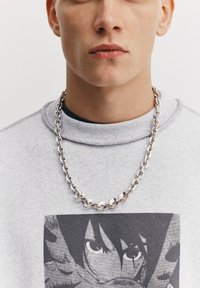 Kædehalskæde i chunky sølv båret over en lysegrå sweatshirt med sort-hvidt grafisk tryk. Nærbillede af nakke og ansigt.