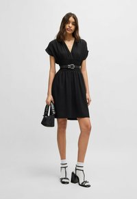 Zwarte jurk met korte mouwen, getailleerde taille en riem. Model houdt een kleine zwarte tas met logo vast. G gecombineerd met gestreepte sokken en zwarte hakken.