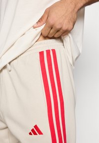 Joggery w jasnobeżowym kolorze z trzema czerwonymi paskami po bokach, elastycznym paskiem w talii i czerwoną logo Adidasa. Widoczna gładka faktura tkaniny.