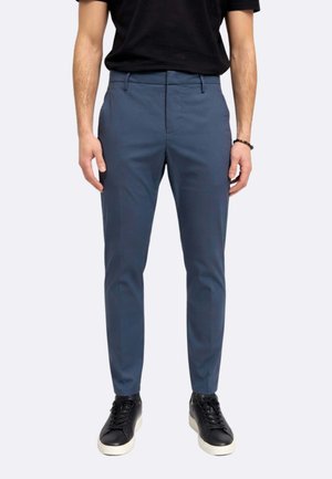 Uomo che indossa pantaloni blu slim fit e scarpe da ginnastica nere con suole bianche, in piedi davanti a uno sfondo chiaro e uniforme.