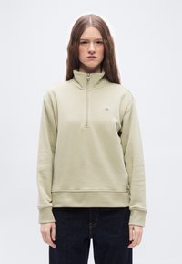 Νεαρή γυναίκα με μακριά μαλλιά, ντυμένη με μπεζ φούτερ με φερμουάρ μέχρι το στήθος και σκουρόχρωμο παντελόνι, στέκεται μπροστά σε ένα απλό λευκό φόντο.