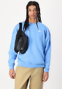 Fekete Nike válltáska, sima textúrával, függőleges cipzárral és logóval. A modellen világoskék pulóver és bézs nadrág van.