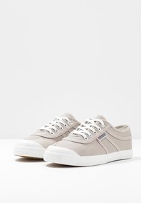 Beige canvas sneakers with white rubber soles, featuring a rounded toe, contrasting side stripes, and white laces.
