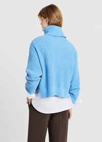 Grobstrick, hellblaues Sweatshirt mit hohem Kragen, cropped Schnitt und Rippstruktur, über einem weißen Hemd und braunen Hosen getragen.
