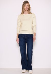Pullover beige chiaro con colletto a girocollo, abbinato a jeans a zampa d'elefante blu scuro con strisce laterali bianche e sneakers bianche.