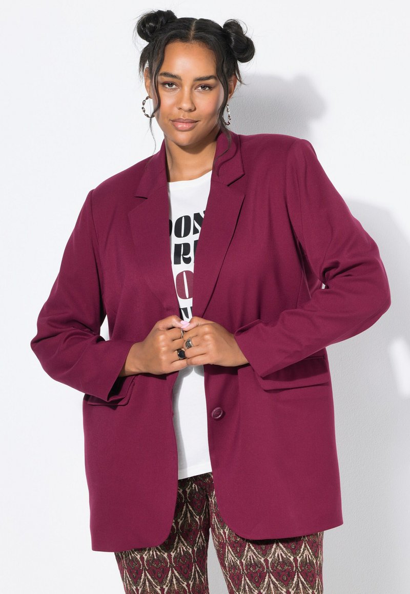 Donna con capelli scuri raccolti in bun, che indossa orecchini a cerchio, una maglietta bianca con grafica, un blazer bordeaux e pantaloni a fantasia, in posa contro un muro bianco.