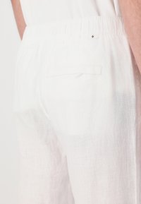 Pantaloni bianchi di lino con vita elastica, dotati di una tasca posteriore singola, tessuto testurizzato e design minimalista senza decorazioni.