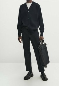 Zwarte geribbelde cardigan met knopen, witte T-shirt, zwarte broek en zwarte leren schoenen. Draagt een donkere tas met schouderriem.