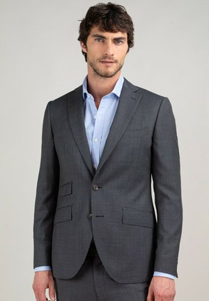Hombre con traje gris oscuro y camisa azul clara, de pie frente a un fondo claro y liso.