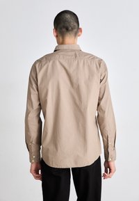 Camisa de manga larga de color marrón claro con cuello abotonado, que presenta una textura suave y detalles sutiles de costura en la yoke trasera.