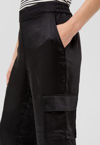 Zwarte satijnen cargo pants met zijzakken en een elastische tailleband. De stof heeft een gladde textuur en een subtiele glans.