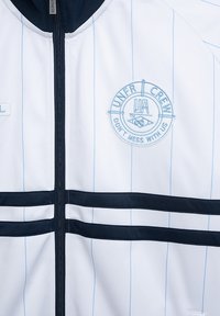 Bijela sportska jakna s svijetloplavim prugama i tamnoplavim detaljima. Ima zatvarač sprijeda i okrugli izvezeni logo s tekstom.